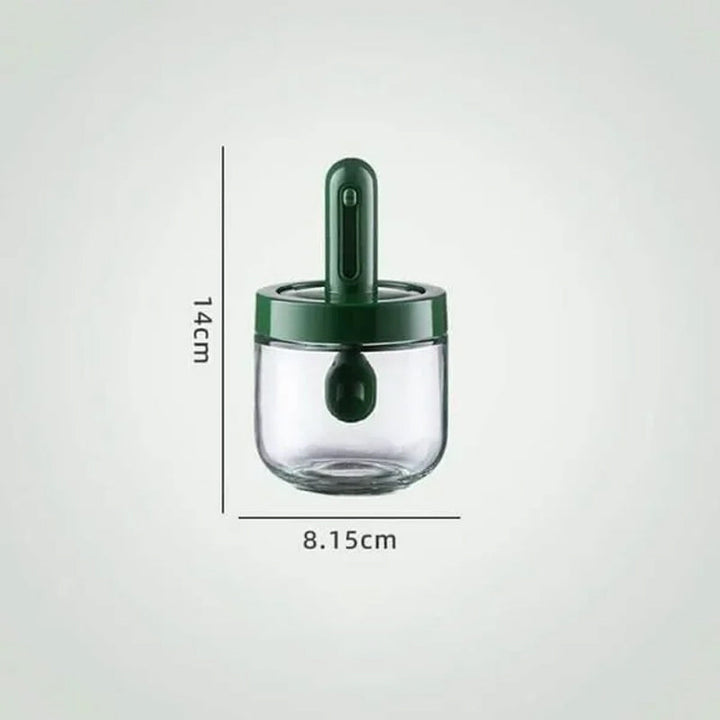 Kryddburk i glas med grönt och vitt skedlock, justerbart för enkel dosering, elegant design för köksanvändning.